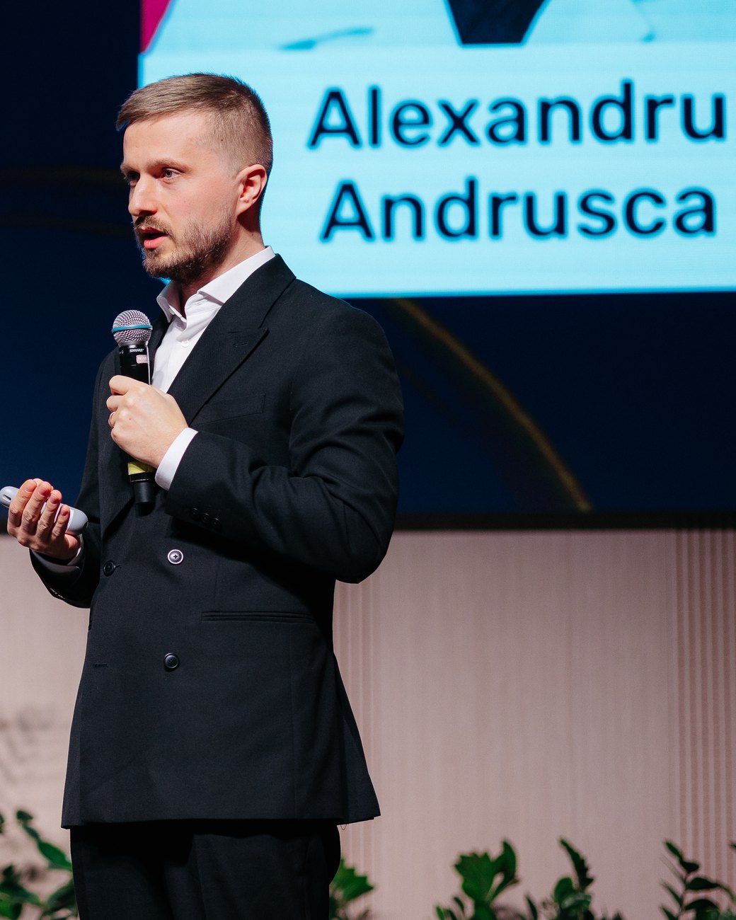 Dr. Alexandru Andrușca vorbind cu microfonul în mână pe scena unei conferințe, cu numele său afișat pe ecranul din spate